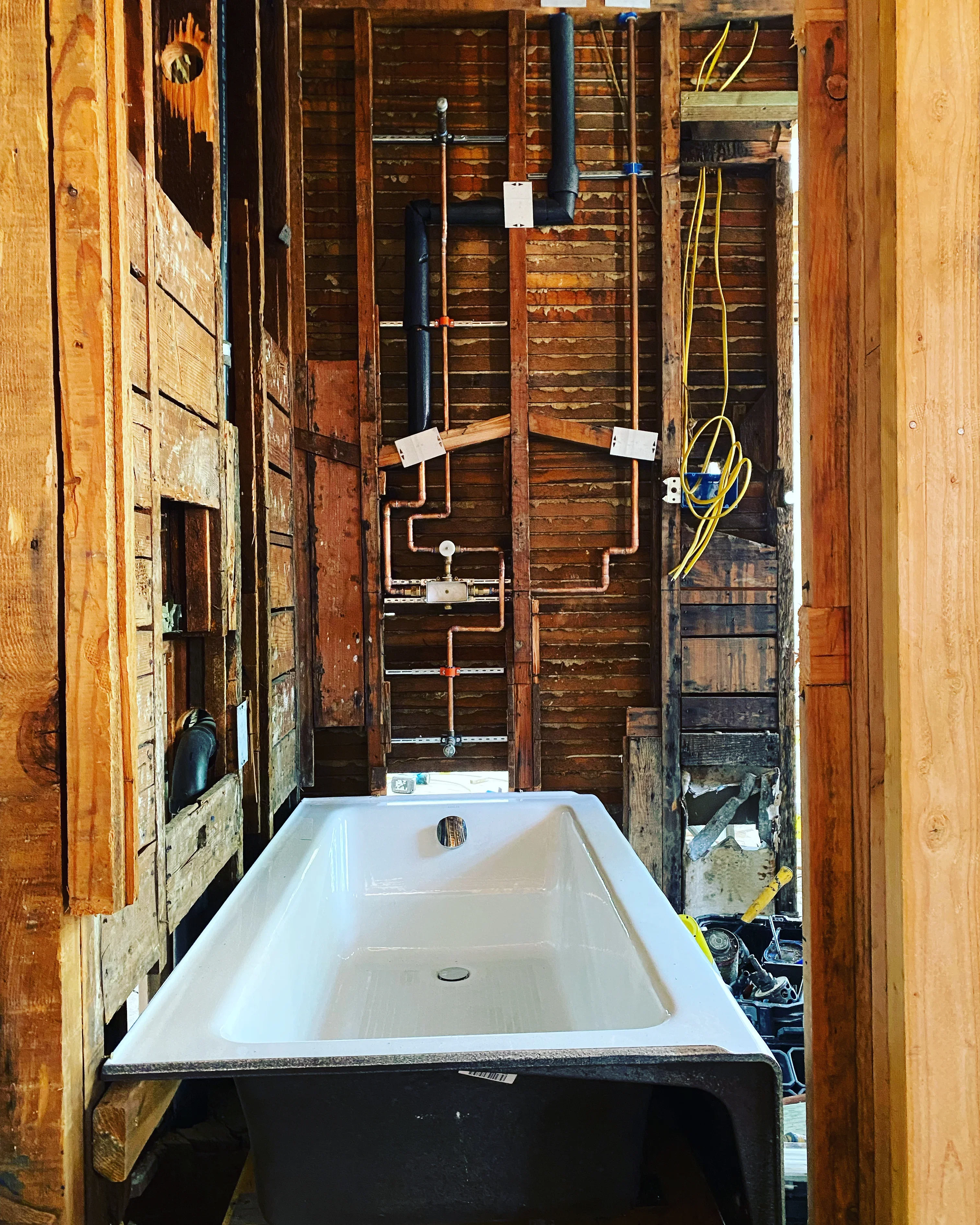 Bathroom plumbing rough-in with white soaking tub, copper and PVC supply lines, and drain piping installed in wooden framed space